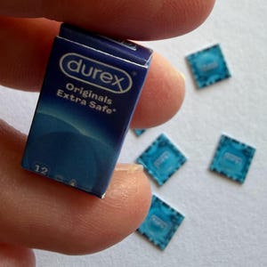 Può includere: Una scatola rettangolare blu in miniatura con l'etichetta Durex Originals Extra Safe e il numero 12 stampato sul davanti. Diverse piccole confezioni quadrate di colore blu, simili a fogli di alluminio, con il logo Durex sono sparse vicino.