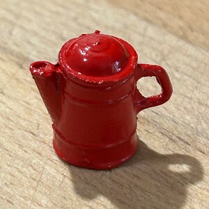 May include: A small, red metal teapot with a rounded lid and a handle. The teapot is sitting on a wooden surface.