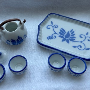 Op de afbeelding: Een miniatuur wit porseleinen theeservies met blauwe bloemmotieven. De set bevat een theepot met een bruin handvat, een deksel, een serveerschaal en drie kleine kopjes. De schaal en kopjes hebben blauwe randen.