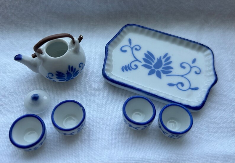 Op de afbeelding: Een miniatuur wit porseleinen theeservies met blauwe bloemmotieven. De set bevat een theepot met een bruin handvat, een deksel, een serveerschaal en drie kleine kopjes. De schaal en kopjes hebben blauwe randen.
