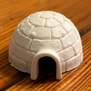 May include: A white plastic model of an igloo with a rounded top and a rectangular entrance. The igloo is designed to look like it is made of bricks.