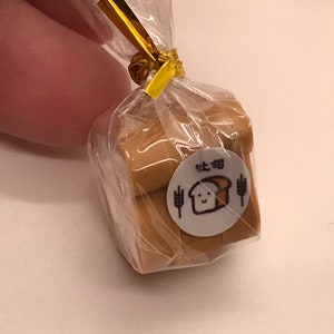 May include: A miniature package of bread slices wrapped in clear cellophane and tied with a gold ribbon. A white sticker with a cartoon bread slice and Japanese text is affixed to the package. The bread is light brown.