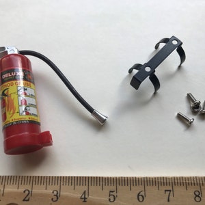 Peut inclure: Un extincteur rouge avec un tuyau noir et un support en métal noir avec des vis. L'extincteur porte les mots "Deluxe" et "Mode d'emploi" imprimés dessus.