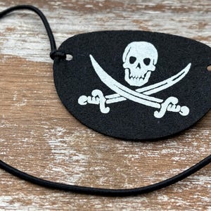 May include: A black felt eye patch featuring a white screen-printed graphic of a skull positioned above two crossed swords. The eye patch is oval-shaped and includes a thin, black elastic cord attached through small holes on each side for securing.