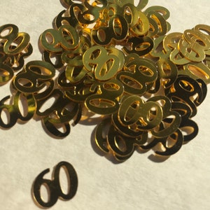 May include: A pile of gold-colored confetti shaped like the number 60. The confetti is scattered on a white surface.
