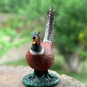 May include: A small, detailed figurine of a pheasant with a long, white-tipped tail. The pheasant is painted in shades of brown, red, and black, with white accents. The figurine is standing on a green base.