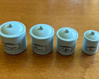 May include: Four small, light blue ceramic canisters with black lids and labels. The labels read "Sugar", "Flour", "Coffee", and "Tea".