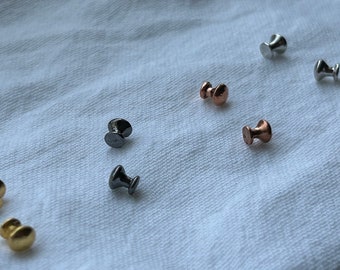 May include: Assortment of small metal knobs in various finishes, including silver, gold, black, and rose gold. The knobs are arranged on a white fabric surface.