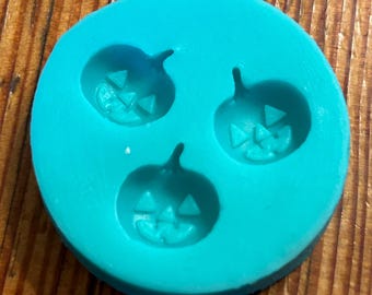 May include: A turquoise silicone mold with three pumpkin-shaped cavities. Each cavity features a jack-o'-lantern face with a triangular nose and eyes.