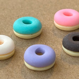 May include: Five miniature donuts with different colored glazes. The donuts are white, pink, light blue, purple, and brown. Each donut has a white frosting ring around the center hole.