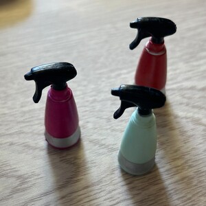 May include: Three small spray bottles in pink, red, and light blue with black spray nozzles. Each bottle has a silver-colored base. The bottles are arranged on a light brown wooden surface.
