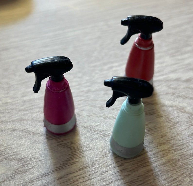 May include: Three small spray bottles in pink, red, and light blue with black spray nozzles. Each bottle has a silver-colored base. The bottles are arranged on a light brown wooden surface.