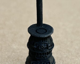 May include: A black cast iron miniature stove with a tall pipe. The stove has a round base with three legs and a decorative design on the front.