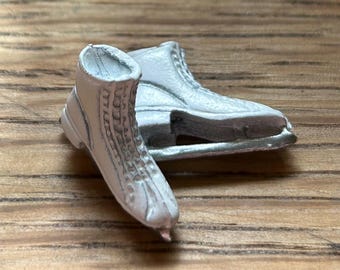 May include: Two white ice skates with silver blades are displayed on a wooden surface. The skates are detailed with laces and a textured design. One skate is upright, while the other lies on its side, showcasing the blades.