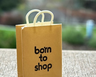 May include: A small brown paper shopping bag with white rope handles. The bag has the text "born to shop" printed on the front in black.