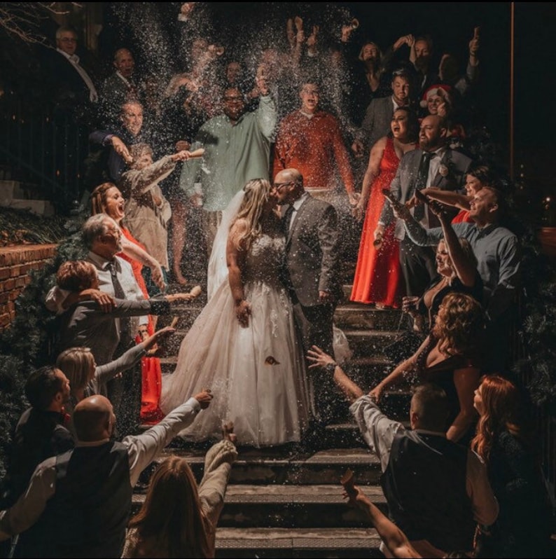 May include: A bride and groom kiss as they walk down a set of stairs, surrounded by guests throwing confetti. The bride is wearing a white wedding dress and the groom is wearing a dark suit. The guests are dressed in various attire, some holding champagne glasses.