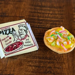 May include: A miniature pizza box with the words "Pizza" and "Made with Real Tomato and Fresh Mozzarella!" printed on it.  Next to the box is a miniature pizza with shrimp, green onions, and red onions on top.