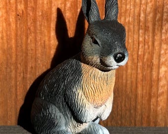 May include: A small, gray and brown rabbit figurine with a white belly. The rabbit is sitting with its ears upright and its eyes closed.