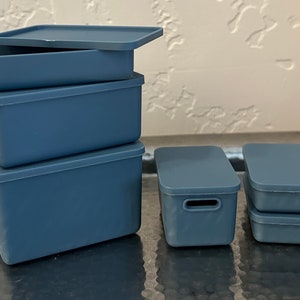 May include: A stack of four blue plastic storage boxes with lids. The top box is open and the other three are closed. The boxes are on a silver surface.
