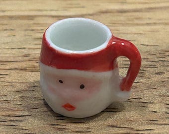Puede incluir: Una pequeña taza de cerámica con forma de Papá Noel. Tiene un gorro rojo y un asa. La cara blanca está adornada con ojos negros y una boca roja.