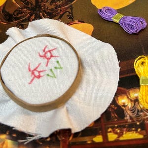 Puede incluir: Un aro de bordado blanco con un diseño floral cosido con hilo rojo y verde. El aro está sobre un fondo de tela blanca.