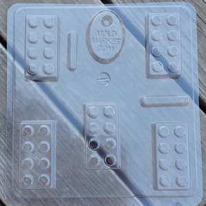 Pode incluir: Forma de plástico transparente com seis cavidades retangulares em forma de blocos Lego. A forma é projetada para criar réplicas de blocos Lego de chocolate ou doces. A forma tem o texto "MOLD MARKET COM" nela.