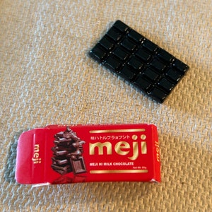 Puede incluir: Un envoltorio rojo y blanco para una barra de chocolate con el texto "meji" y "MEJI HI MILK CHOCOLATE" impreso en él. El envoltorio muestra una pila de trozos de chocolate. También se muestra una barra de chocolate negra en la imagen.