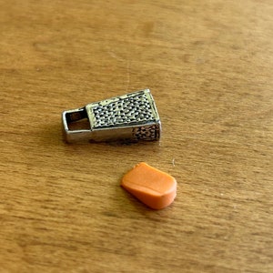 Op de afbeelding: Een zilveren rits-trekker met een getextureerd oppervlak en een klein oranje rubberen stuk.
