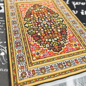 May include: A miniature, rectangular rug with a detailed floral pattern. The rug features a central design in black, red, and yellow, surrounded by a yellow border with floral accents. The edges are finished with a white fringe.