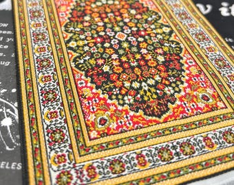 May include: A miniature, rectangular rug with a detailed floral pattern. The rug features a central design in black, red, and yellow, surrounded by a yellow border with floral accents. The edges are finished with a white fringe.