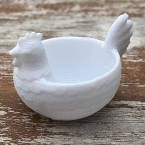 May include: A white 3D printed bowl shaped like a nesting hen. The bowl features a sculpted chicken head on one side and a raised tail feather design on the opposite side, with a textured, layered surface finish throughout the entire object.