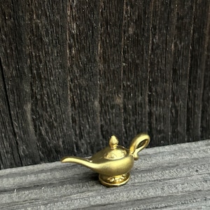 Puede incluir: Una lámpara de genio dorada con detalles intrincados. La lámpara tiene un diseño tradicional con un pico curvo y una base redonda.