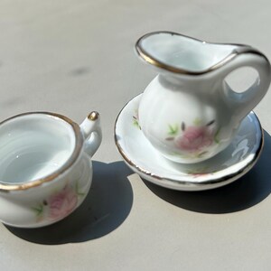 May include: A miniature white porcelain pitcher with a gold rim and floral design, on a matching saucer.  Next to it is a miniature white porcelain chamber pot with a gold rim and floral design.