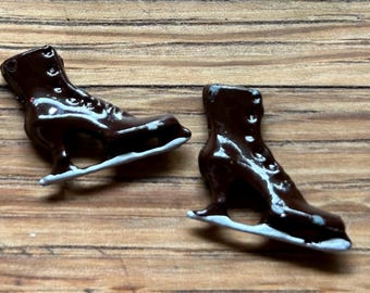 May include: Two brown ice skate charms with white blades. The skates have a shiny finish and are detailed with laces and a strap.
