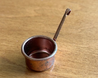 May include: A small, copper-colored saucepan with a long handle and a hook at the end. The miniature pot has a rounded bowl and is designed for measuring or serving small portions. The metal has a slightly aged appearance.