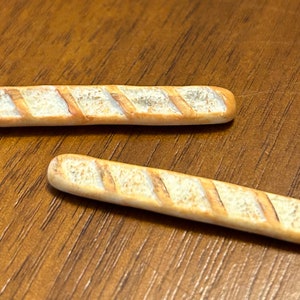 May include: Two miniature, light brown, baguette-shaped loaves of bread on a wooden surface.