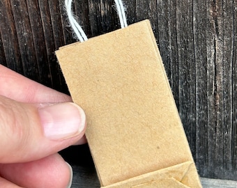 May include: A small brown paper bag with white string handles. The bag is folded and sitting on a wooden surface.