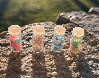 May include: Four small glass jars filled with colorful sprinkles. The jars have white lids and are arranged in a row on a brown, textured surface.