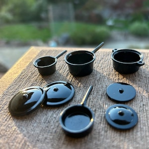 May include: A set of miniature black cookware, including pots, pans, and lids. The cookware is made of metal and has a shiny finish.