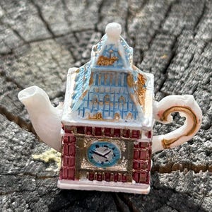 May include: A white ceramic teapot shaped like a clock tower with a blue and gold roof. The teapot has a red brick base and a clock face with gold hands.