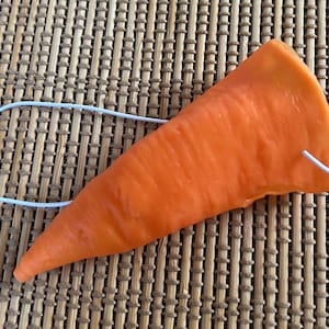 May include: An orange carrot nose mask with a white elastic band. The mask is shaped like a carrot and is designed to be worn on the face. The mask is made of a soft material.