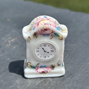 May include: A small, white porcelain clock with a floral design. The clock face has Roman numerals and hands pointing to approximately 2:10. The clock is decorated with pink, red, yellow, and green floral patterns.