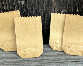 May include: A collection of six small, folded brown paper bags. The bags have a rectangular shape with a zig-zag top edge and a folded bottom. They are arranged on a wooden surface with a dark wood background.