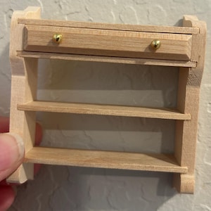 May include: A miniature wooden shelf unit with a drawer and two shelves. The drawer has two small gold knobs. The wood is light-colored and appears unfinished. The unit is designed for a dollhouse or other miniature setting.