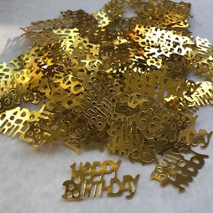 May include: Gold confetti with the words "Happy Birthday" cut out. The confetti is scattered on a white surface.