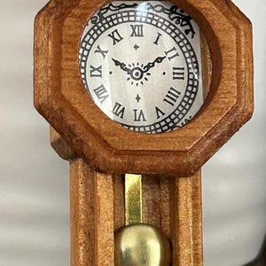 Puede incluir: Pequeño reloj de madera con acabado marrón claro. La esfera tiene números romanos negros y detalles ornamentales. Un péndulo dorado cuelga debajo. El reloj es un artículo decorativo, ideal para coleccionistas.