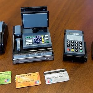 May include: A collection of miniature point-of-sale (POS) system components, including a receipt printer, cash register, payment terminal, and barcode scanner. Three debit cards are also present. The items are black, gray, and silver.
