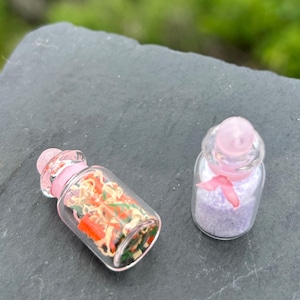 May include: Two small glass bottles with pink accents. One bottle contains colorful, dried ingredients, while the other holds lavender-colored granules and a pink ribbon. The bottles are displayed on a dark gray surface.