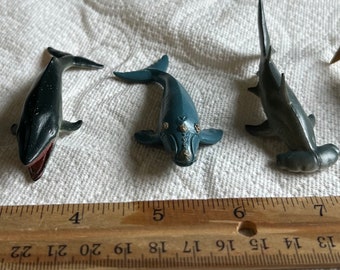 May include: A set of six plastic toy sea creatures, including a whale shark, a humpback whale, a right whale, a hammerhead shark, a tiger shark, and a whale. The toys are arranged on a white surface with a wooden ruler for scale.