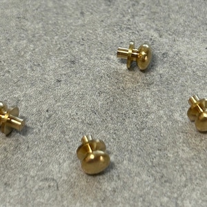 May include: Four gold-toned metal knobs with a round top and a small post on the bottom. The knobs are arranged in a circle on a gray surface.